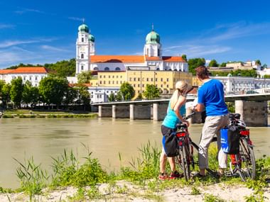 Zwei Radfahrer mit Packtaschen stehen am Inn und blicken auf Passaus Altstadt mit Domtürmen und Brücke unter blauem Himmel.