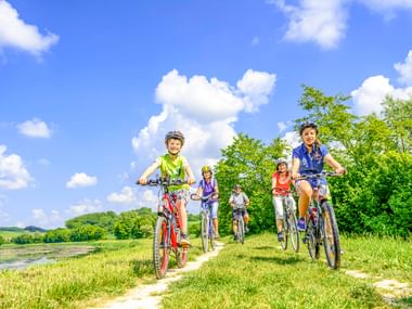 Five cyclists with helmets riding on a grass path beside a river under blue sky with white clouds. Green trees and hills in background.
