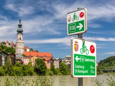 Tauernradweg R3A Radwegeschild mit Richtungsangaben nach Salzburg und St. Radegund, mit Burghausens Kirchturm und bunten Gebäuden im Hintergrund.