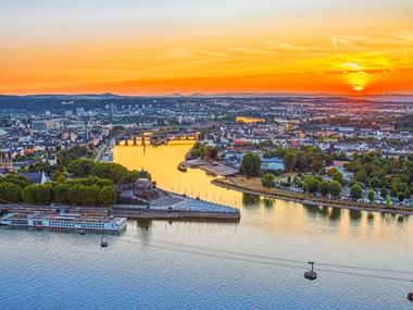 Luftaufnahme des Deutschen Ecks in Koblenz bei Sonnenuntergang mit goldenem Licht über dem Zusammenfluss der Flüsse, Seilbahnen und Stadtbild.