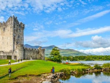 Ross Castle, ein mittelalterliches Steinturmhaus mit Zinnen, steht auf grünen Rasenflächen neben einem See in Killarney mit Bergen im Hintergrund.