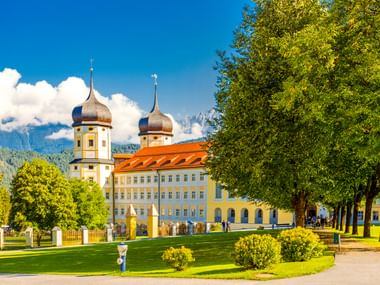 Stift Stams monastery in Tirol with yellow baroque building, twin onion-domed towers, orange roof, green lawn and tree-lined path under blue sky.
