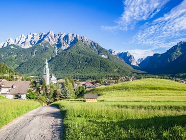 Weg durch grüne Wiesen nach Toblach mit weißem Kirchturm, umgeben von bewaldeten Bergen und markanten Dolomitengipfeln.