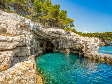Weiße Kalksteinklippen mit natürlicher Höhle zum türkisfarbenen Wasser bei Pula, Kroatien. Grüne Bäume krönen die Felsformation unter blauem Himmel.