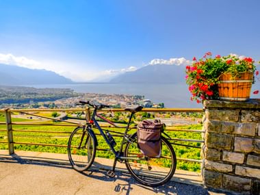 Fahrrad mit Taschen neben Steinpfeiler mit Blumen, mit Blick auf Genfer See und Berge bei La Tour-de-Peilz.