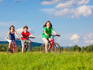 Drei Kinder fahren auf roten Fahrrädern durch eine grüne Wiese unter blauem Himmel. Bewaldete Hügel im Hintergrund.