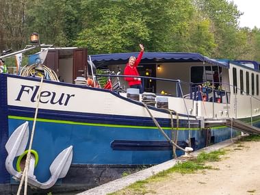 Flusskreuzfahrtschiff MS Fleur am Kanalufer vertäut. Eine Person in roter Jacke winkt vom Oberdeck. Fahrräder sind an Deck sichtbar.