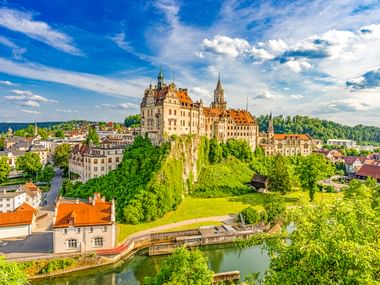 Hohenzollernschloss auf einem Hügel in Sigmaringen, umgeben von üppigem Grün und der Stadt darunter, mit einem Fluss im Vordergrund unter blauem Himmel.