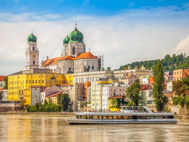Der Passauer Dom St. Stephan mit grünen Zwiebeltürmen am Donauufer. Ein Kreuzfahrtschiff fährt an bunten historischen Gebäuden vorbei.