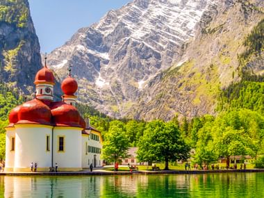 St. Bartholomä Kirche mit roten Zwiebeltürmen am Königssee, umgeben von grünen Wiesen und dramatischen Berggipfeln.