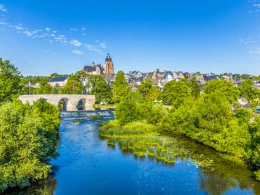 Blick auf Wetzlarer Altstadt mit historischem Kirchturm und Steinbrücke über die Lahn, umgeben von grünen Bäumen unter blauem Himmel.