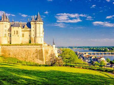 Schloss Saumur mit weißen Steinmauern und Türmen auf einem Hügel, mit Blick auf die Loire und historische Brücke unter blauem Himmel.