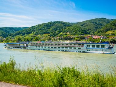 Flusskreuzfahrtschiff MS Swiss Crown fährt auf der Donau mit grünen Hügeln und einem Dorf im Hintergrund unter blauem Himmel.