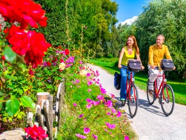 Zwei Radfahrer auf roten Rädern fahren auf einem asphaltierten Weg in Ahrenshoop, umgeben von bunten Blumen und grünen Bäumen.