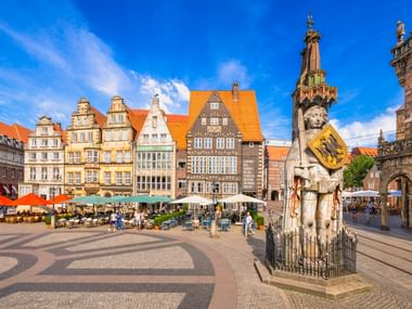 Bremer Marktplatz mit der Rolandstatue vor bunten historischen Gebäuden mit orangen Dächern. Straßencafés mit Sonnenschirmen säumen den Platz.