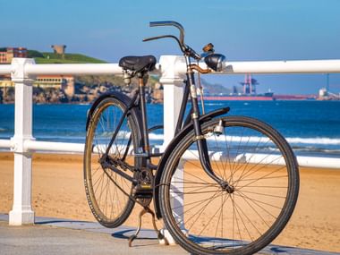 Schwarzes Fahrrad lehnt an weißem Geländer am Strand von Gijón. Sandstrand und blaues Meer mit Frachtschiff im Hintergrund.