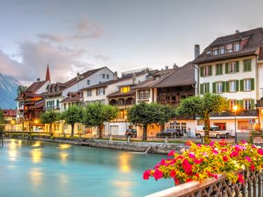 Interlaken Flussufer bei Abenddämmerung mit traditionellen Schweizer Gebäuden, türkisfarbenem Aare-Fluss und leuchtenden Blumen im Vordergrund.