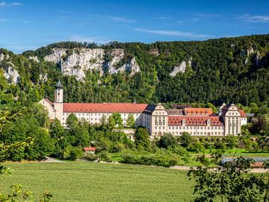Kloster Beuron mit Kirchturm und rotgedeckten Gebäuden im grünen Tal unterhalb markanter weißer Kalksteinfelsen und Wald.