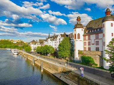 Weiße Alte Burg mit runden Türmen am Fluss in Koblenz. Uferpromenade mit Bäumen und weißen Gebäuden unter blauem Himmel.