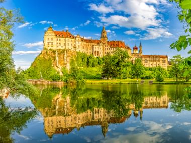 Hohenzollernschloss in Sigmaringen auf einer Klippe über der Donau, mit üppigen grünen Bäumen und perfekter Spiegelung im ruhigen Wasser.