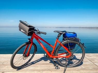 Rotes 30-Gang-Unisex-Tourenrad mit Lenkertasche und blauen Gepäcktaschen auf Holzsteg an ruhigem See unter blauem Himmel.