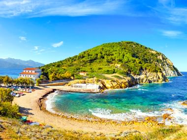 Geschwungener Strand mit türkisfarbenem Wasser an der Enfola-Halbinsel in Portoferraio, Elba. Ein grüner bewaldeter Hügel erhebt sich aus dem Meer, links ist ein weißes Gebäude sichtbar.