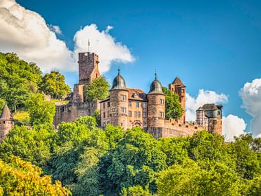 Burg Wertheim mit Steintürmen und Türmchen thront auf einem grünen bewaldeten Hügel unter blauem Himmel mit weißen Wolken.