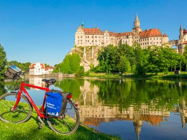 Rotes Tourenrad mit blauen Taschen am Flussufer mit Schloss Sigmaringen, das sich in der Donau unter blauem Himmel spiegelt.