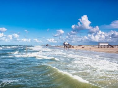 Breiter Sandstrand in St. Peter-Ording mit Wellen, Pfahlbauten in der Ferne und dramatischen Wolken am blauen Himmel.