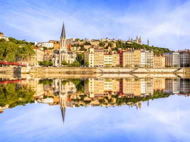 Historische Gebäude der Lyoner Altstadt mit Kirchturm, spiegeln sich im ruhigen Wasser der Saône unter blauem Himmel.