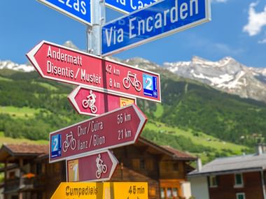 Radweg-Wegweiser in Sumvitg mit Richtungen nach Andermatt, Disentis, Chur und Ilanz mit Entfernungen. Alpenberge und grüne Wiesen im Hintergrund.