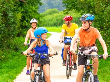 Familie mit fünf Personen radelt auf asphaltiertem Weg entlang des Lahn-Radwegs. Alle tragen Helme und bunte Kleidung, umgeben von grünen Bäumen.