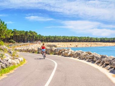 Cyclist in red shirt riding along paved coastal path with rocky shoreline, blue water, sandy beach and pine forest under sunny sky near La Palmyre.