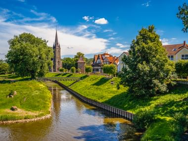Kanal mit grasbewachsenen Ufern im alten Hafen von Rinteln. Kirchturm und Fachwerkhäuser zwischen grünen Bäumen unter blauem Himmel.