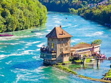 Schlössli Wörth auf einem Felsen im türkisfarbenen Rhein bei Schaffhausen. Besucher auf Aussichtsplattform, üppige grüne Wälder am Flussufer.