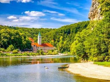 Kloster Weltenburg mit weißem Glockenturm und roten Dächern am Donauufer. Kajakfahrer paddeln nahe einem Sandstrand, umgeben von grünen Hügeln.