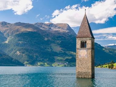 Historischer Kirchturm im Reschensee mit Bergen im Hintergrund unter blauem Himmel mit weißen Wolken.
