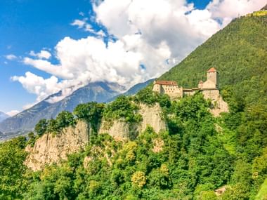 Schloss Tirol thront auf einer Felsklippe umgeben von grünem Wald, mit Bergen und weißen Wolken im Hintergrund unter blauem Himmel.