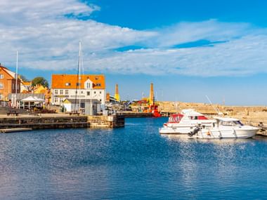 Hafen von Allinge auf Bornholm mit weißen Booten im ruhigen blauen Wasser. Gelbe und weiße Gebäude säumen die Uferpromenade unter blauem Himmel.