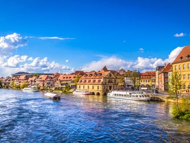 Bamberger Hafen mit bunten historischen Gebäuden entlang des Ufers. Passagierschiffe liegen am Kai unter blauem Himmel mit weißen Wolken.
