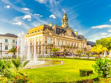 Tours City Hall, an ornate Renaissance-style building with clock tower, behind manicured lawns, fountains, and palm trees under blue sky.