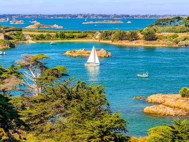 Scenic view of Île de Bréhat showing turquoise waters with a white sailboat, rocky islands, green pine trees, and coastal houses under clear sky.