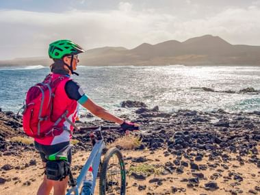 Cyclist with green helmet and pink backpack standing with bike on volcanic rocky beach in Lanzarote, overlooking ocean and mountains.