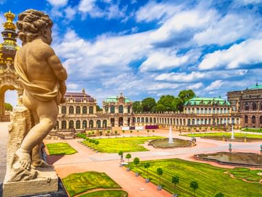 Barocke Statue mit Blick auf den Zwinger-Innenhof in Dresden mit prächtigen Gebäuden, gepflegten Gärten, Springbrunnen und blauem Himmel.