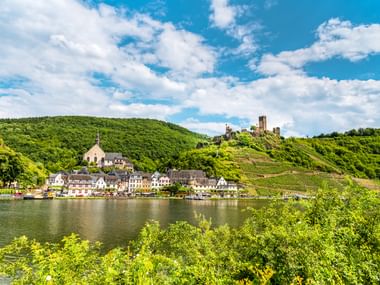 Dorf Beilstein an der Mosel mit Burgruine Metternich auf einem weinbewachsenen Hang unter blauem Himmel mit weißen Wolken.