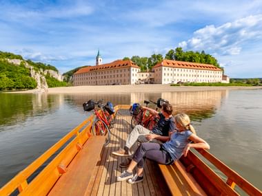 Radfahrer auf Holzboot nähern sich Kloster Weltenburg an der Donau. Das historische Kloster mit roten Ziegeldächern liegt neben Kalksteinfelsen.