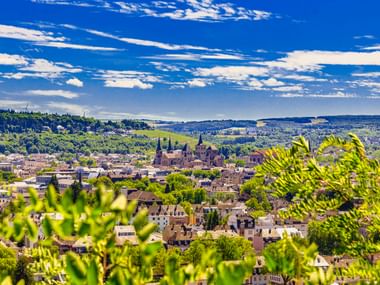Panoramablick auf Trier mit Domtürmen in der Mitte, umgeben von grünen Bäumen und Hügeln unter blauem Himmel mit Wolken.