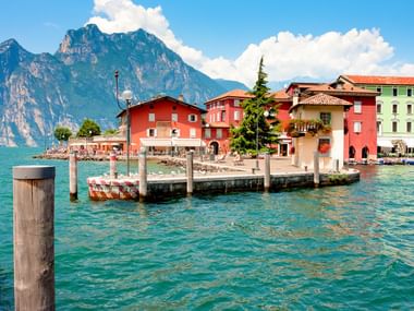 Bunte Gebäude in Torbole am Gardasee mit hölzernen Stegpfosten im türkisfarbenen Wasser. Berge erheben sich im Hintergrund unter blauem Himmel.