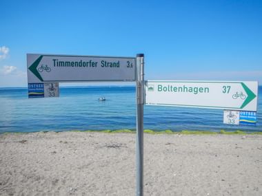 Wegweiser für den Ostsee-Radweg am Sandstrand. Schilder zeigen nach Timmendorfer Strand (3,6 km) und Boltenhagen (37 km).