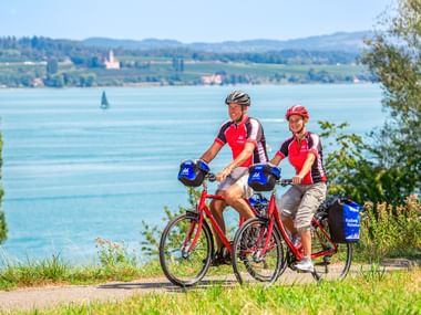 Zwei Radfahrer mit roten Helmen und Trikots auf Tourenrädern mit blauen Taschen, pausierend auf einem Weg mit Blick auf den Bodensee bei Konstanz.